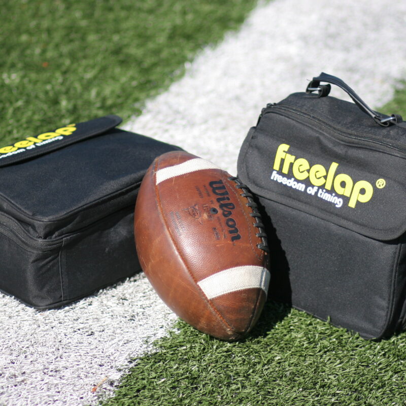 A football, emblematic of sprint-based football action, rests on a white sideline of a grassy field, flanked by two sleek black Freela bags.