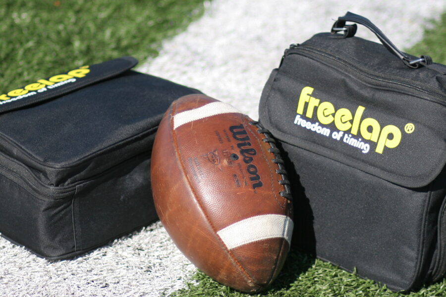 A football, emblematic of sprint-based football action, rests on a white sideline of a grassy field, flanked by two sleek black Freela bags.