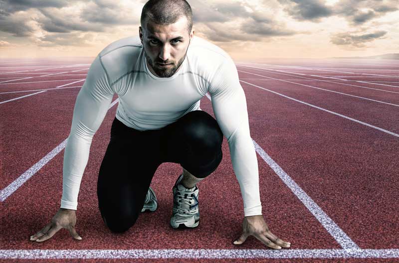 A man in athletic wear crouches in a sprinters stance on a red track, ready to start running. The sky above is cloudy, creating a dramatic backdrop that reflects his focus on potenciación and performance.