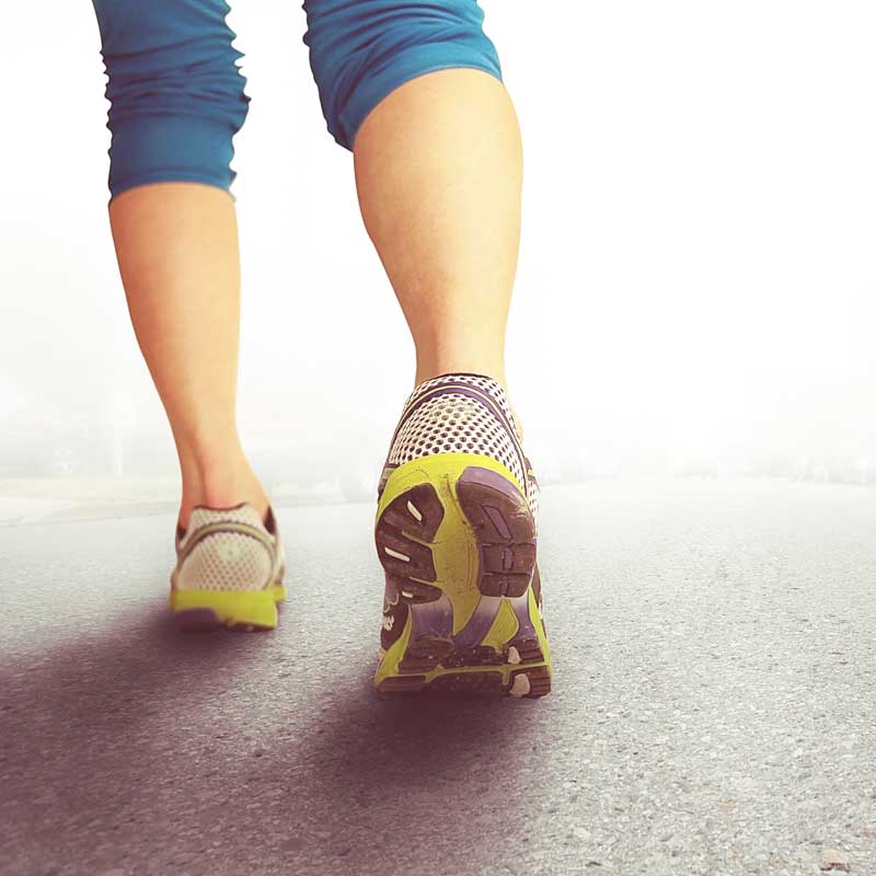 A person wearing blue leggings and sneakers with lime green soles, featuring an ankle rocker design, is walking on a paved surface. The scene is captured from behind at ground level, with the background bright and hazy.