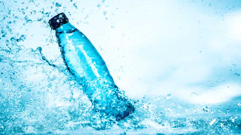 A blue plastic water bottle with a black cap splashes into clear water, representing the perfect hydration choice for competition day, creating dynamic ripples and droplets against a light blue background.