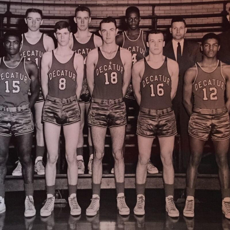 A vintage black and white photo of a basketball team wearing Decatur jerseys. The group consists of eleven players and two coaches. They pose in two rows inside a gym. The players are in uniform with numbers 7, 15, 8, 18, 16, 12, and 17 visible.