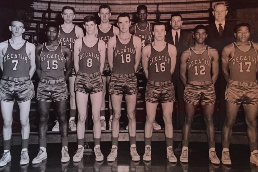 A vintage black and white photo of a basketball team wearing Decatur jerseys. The group consists of eleven players and two coaches. They pose in two rows inside a gym. The players are in uniform with numbers 7, 15, 8, 18, 16, 12, and 17 visible.