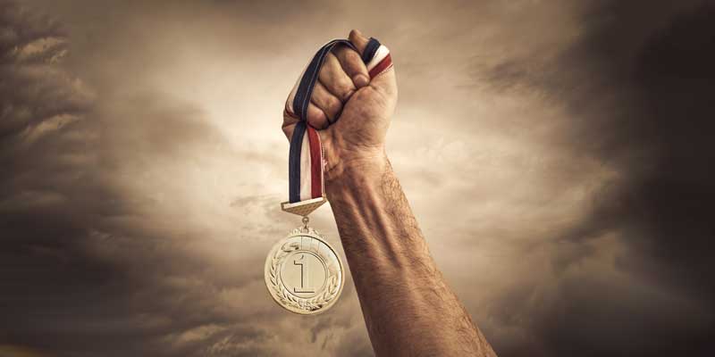 A hand triumphantly holds up a gold medal with a blue, white, and red ribbon against a dramatic cloudy sky background.