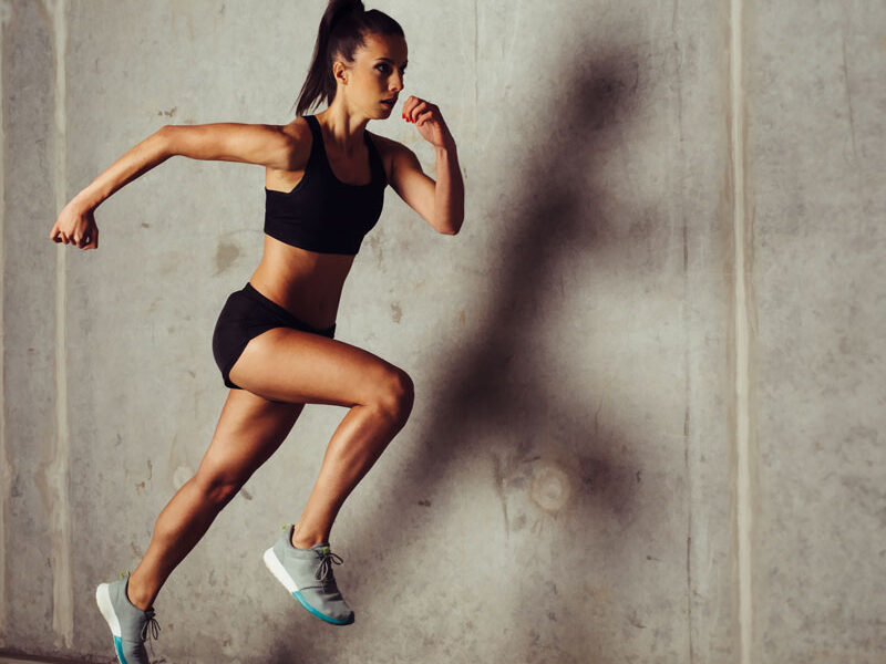 A person in athletic wear leaps in mid-air against a concrete wall backdrop. The individual has their hair in a ponytail and is wearing a black sports bra, black shorts, and white sneakers, showcasing energy and strength.