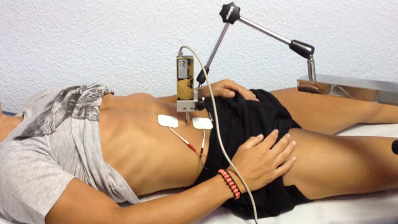 A person is lying on their back with electrodes placed on their abdomen, connected to a machine via wires for a tensiomyography procedure. They are wearing a gray shirt and black shorts, with their right hand resting on their stomach, suggesting the precision of this medical assessment.