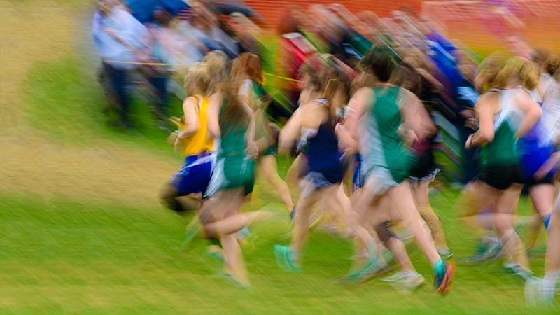 A blurred image of a group of people running on a grassy field. The runners are wearing athletic gear in various colors, including green, blue, and yellow. The motion blur conveys speed and movement. Spectators stand in the background.