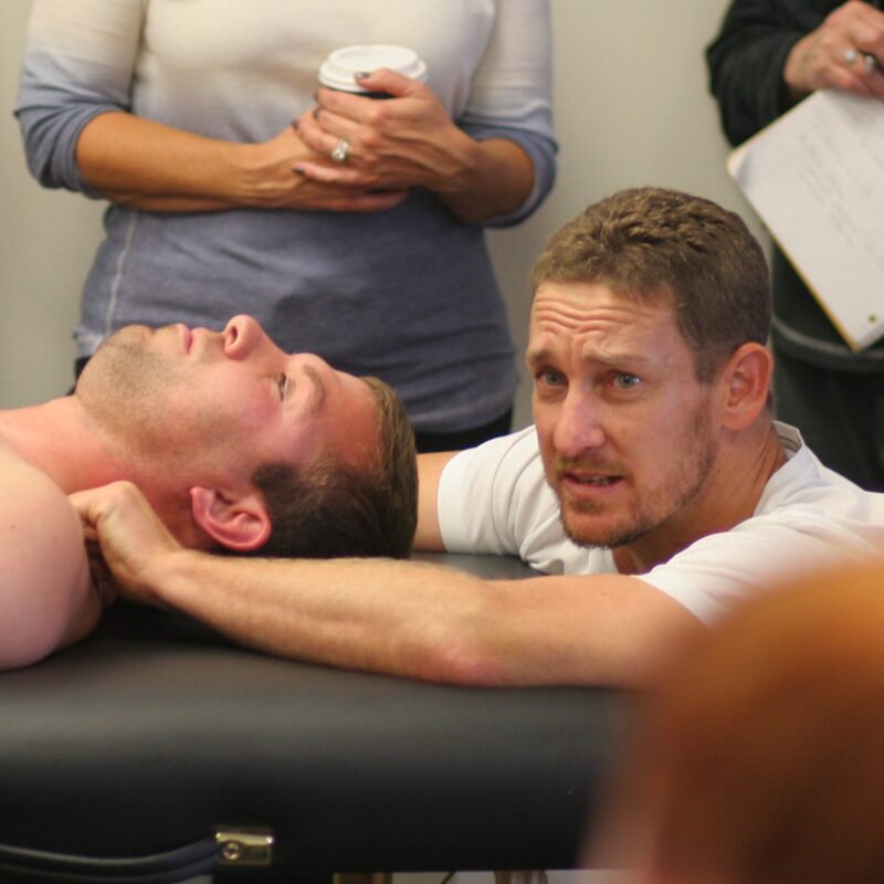A person lying on a massage table while another individual demonstrates a neck massage technique. Several people are observing, some holding notes and a cup. The setting appears educational or instructional.