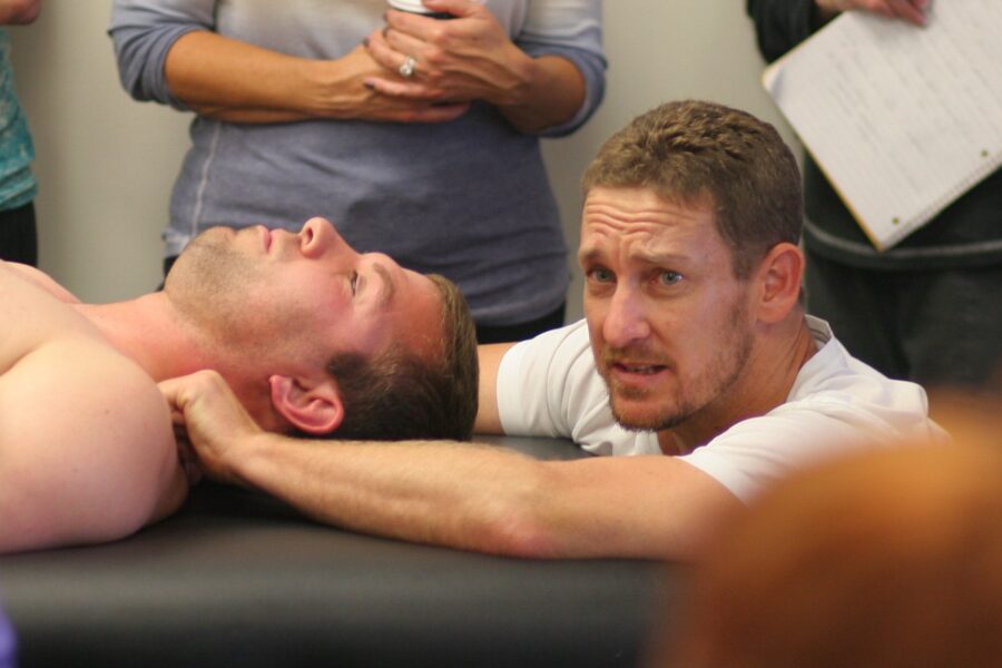 A person lying on a massage table while another individual demonstrates a neck massage technique. Several people are observing, some holding notes and a cup. The setting appears educational or instructional.