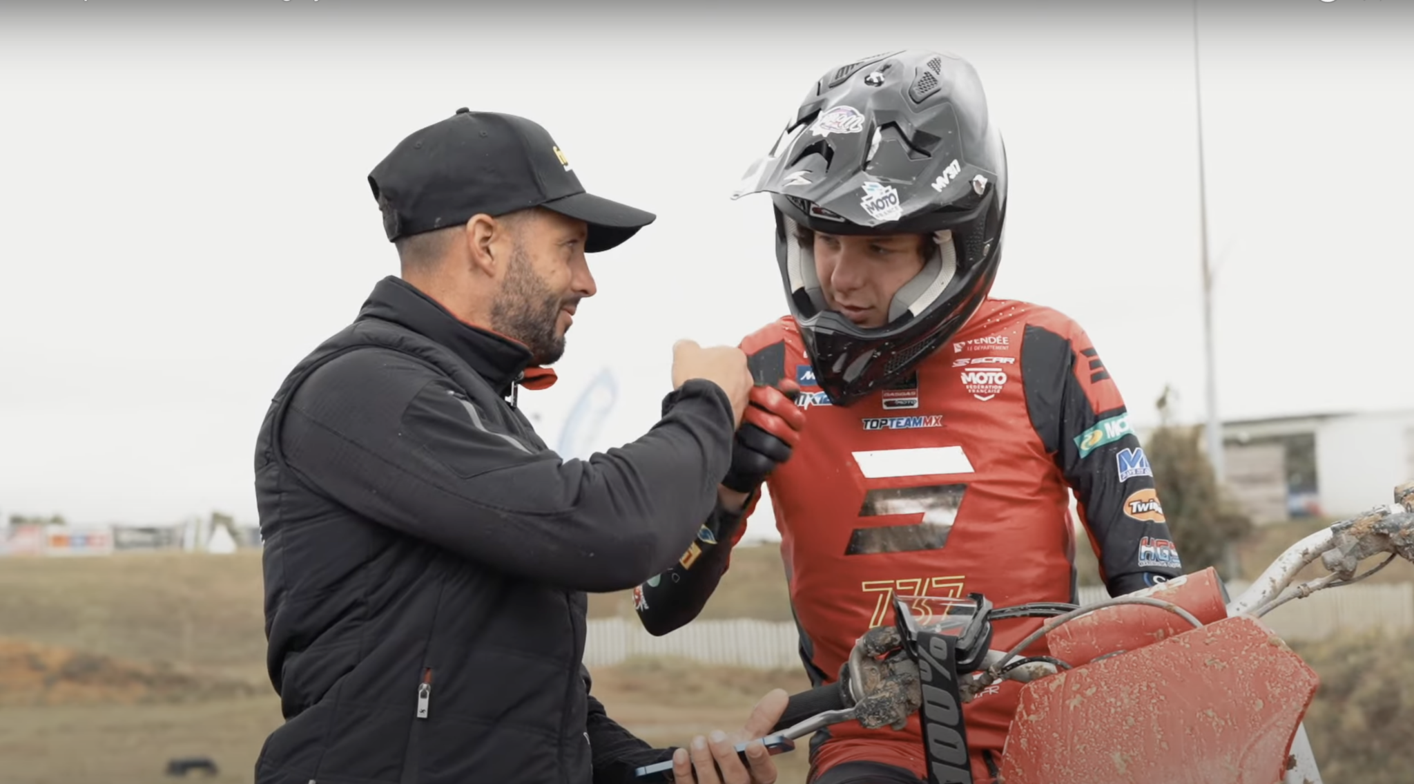 A motocross rider in a red outfit and helmet sits on a dirt bike, receiving a handshake from a person in a black cap and jacket. They are outdoors with a blurred background of the track and equipment.