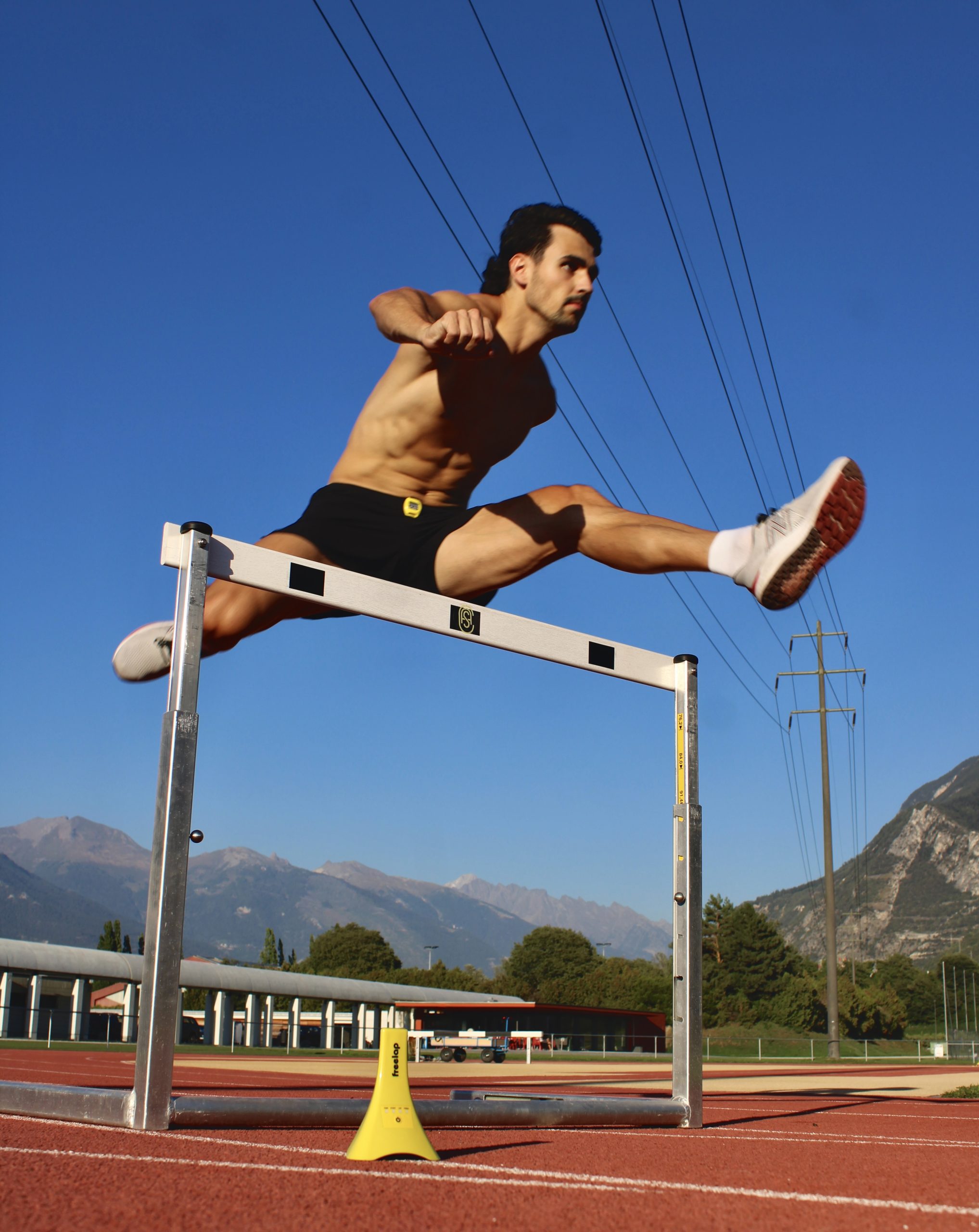 A male athlete in mid-air, wearing black shorts and white shoes, clears a hurdle on a track. He is on an outdoor field with mountains and power lines in the background under a clear blue sky.