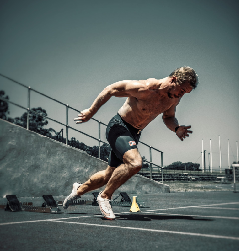 A shirtless man in athletic shorts and sneakers is sprinting on a track, his powerful start captured by the electric timing system. The background of stadium seating and a clear sky suggests an outdoor athletic facility, perfect for showcasing high-speed performance.