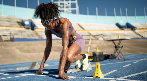 A runner in athletic gear crouches at the starting line on a blue track, preparing to race. The stadium is empty with blue and yellow seats in the background. A cone and a table with equipment are nearby.